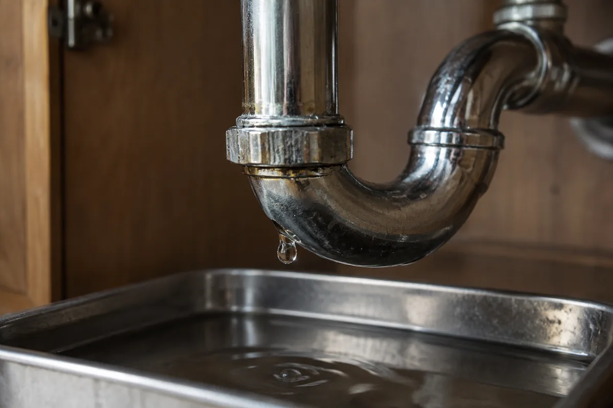 Water droplet beneath a metal sink drainpipe above a shallow catching tray.
